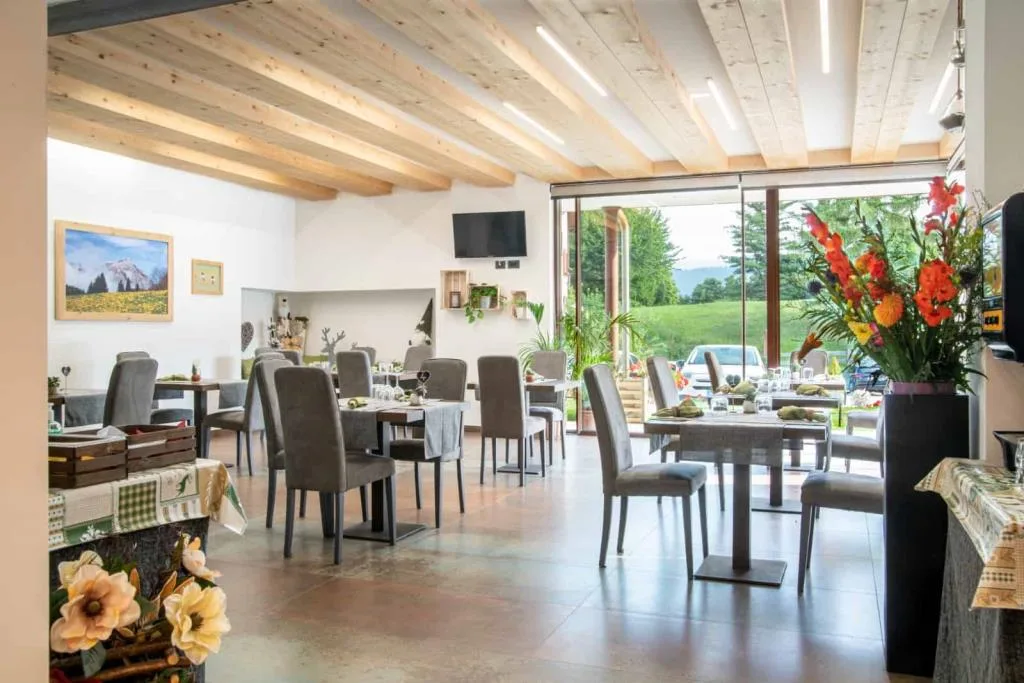 Sala ristorante luminosa con soffitto in travi di legno, ampie vetrate e composizione floreale