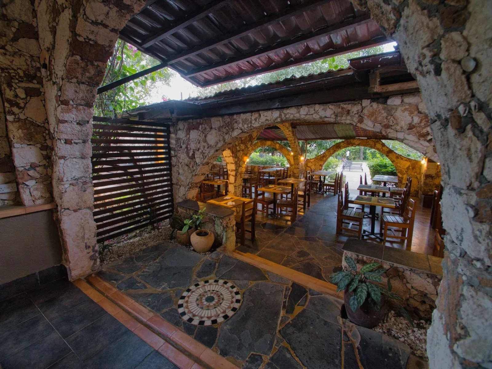 Area ristorante esterna con archi in pietra, tavoli in legno e pavimento decorato a mosaico
